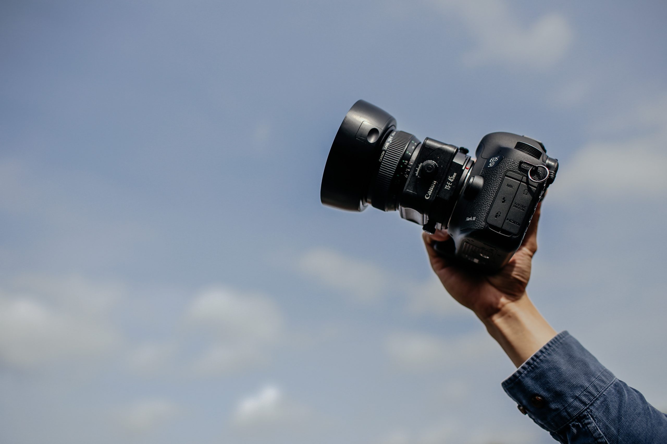 A blue sky background with a hand holding up a black DSLR camera