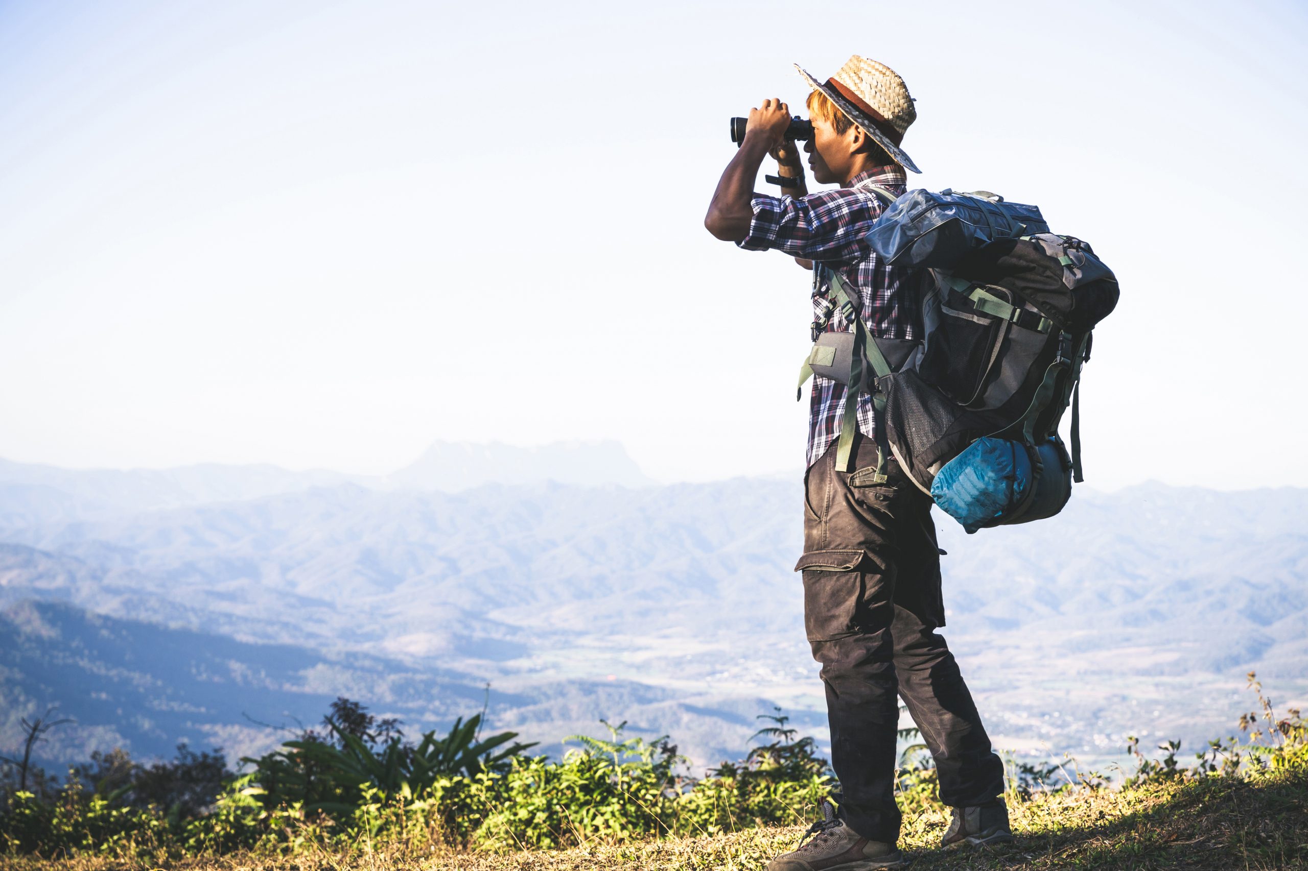 A man is standing on a mountain top, looking through binoculars. He is wearing a large backpack and a hat.