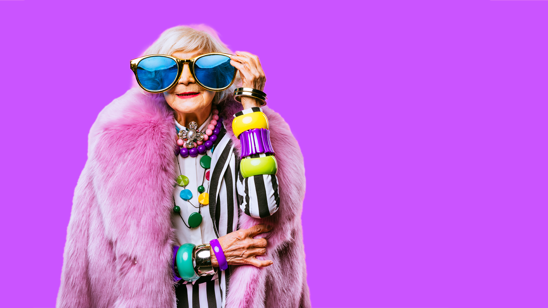 An elderly woman wearing ridiculously oversized sunglasses, extravagant jewellery and a pink fur coat poses confidently